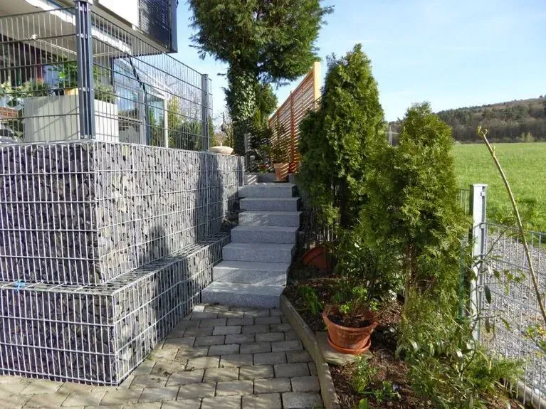 Eine Treppe führt hinauf zu einem Haus, das von Pflanzen und einem Zaun umgeben ist.