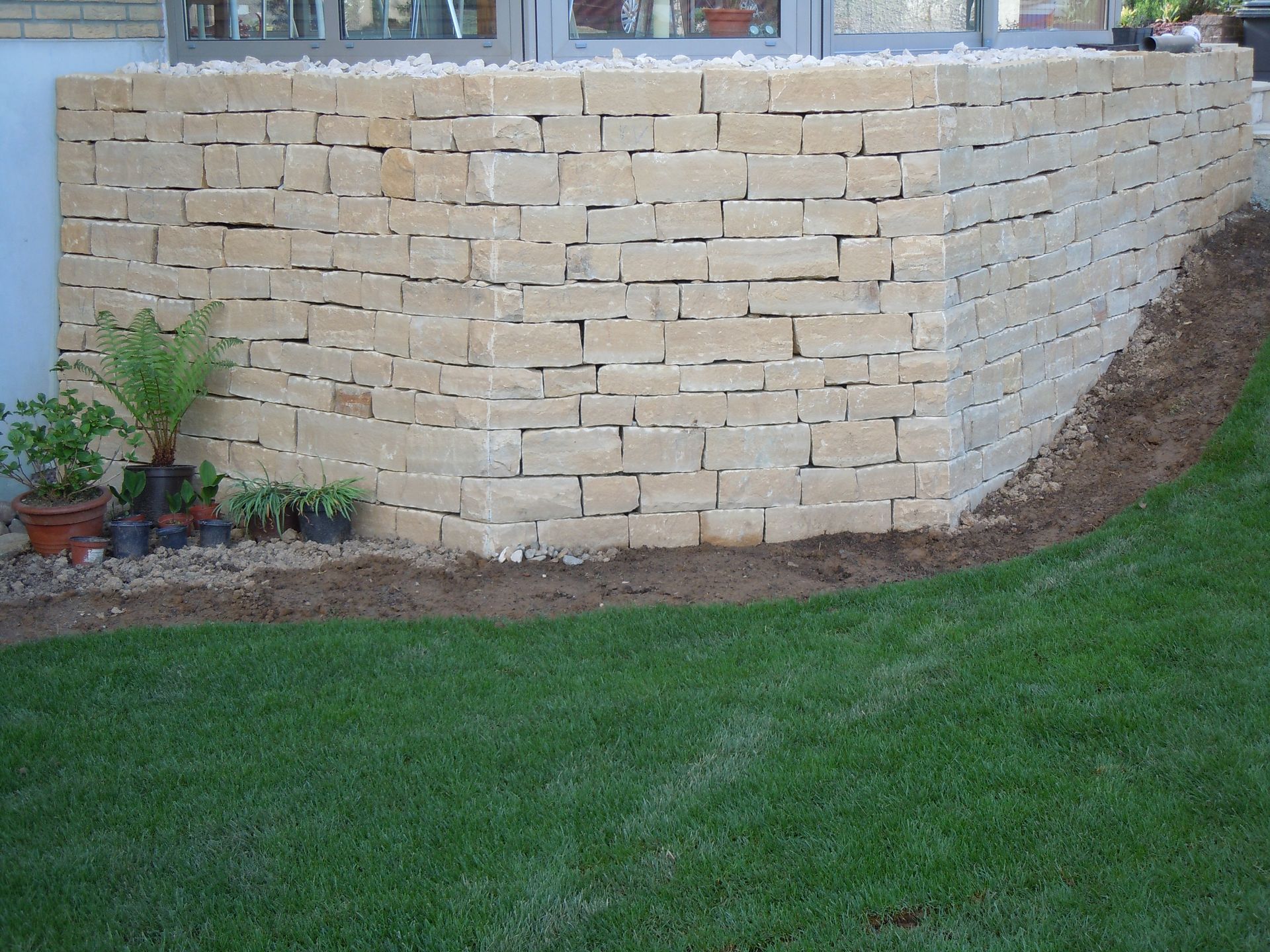 Steinmauer vor einem Gebäude, im Vordergrund Grün und eine Rasenfläche.