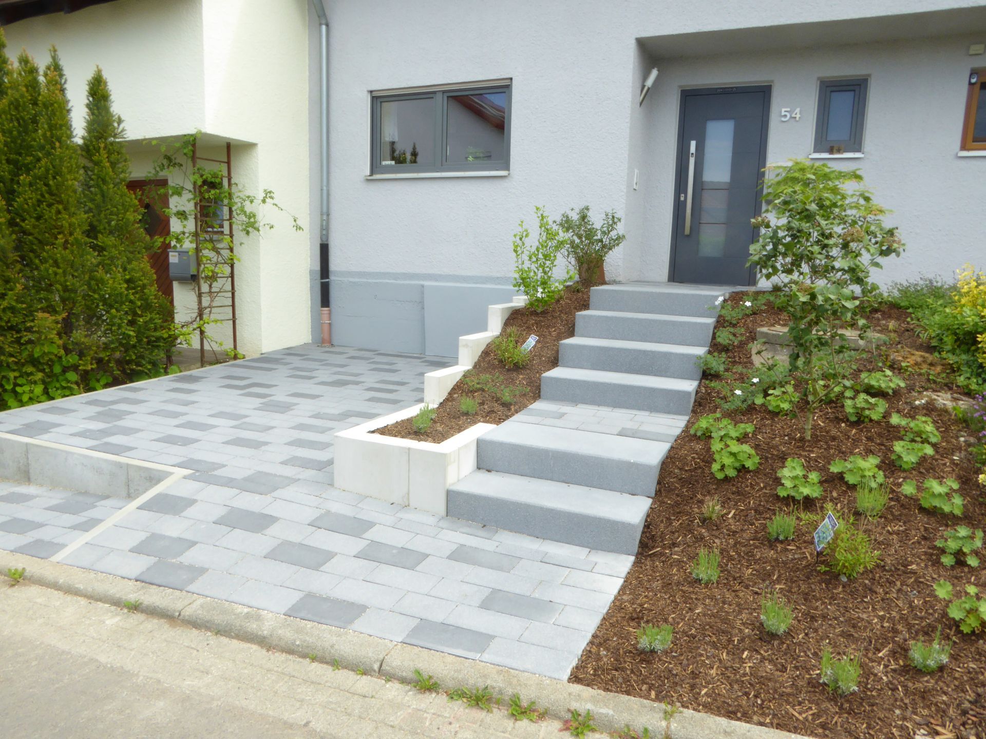 Graue Stufen führen zu einer grauen Tür. Pflanzen und Mulch säumen die Stufen und den Gehweg.