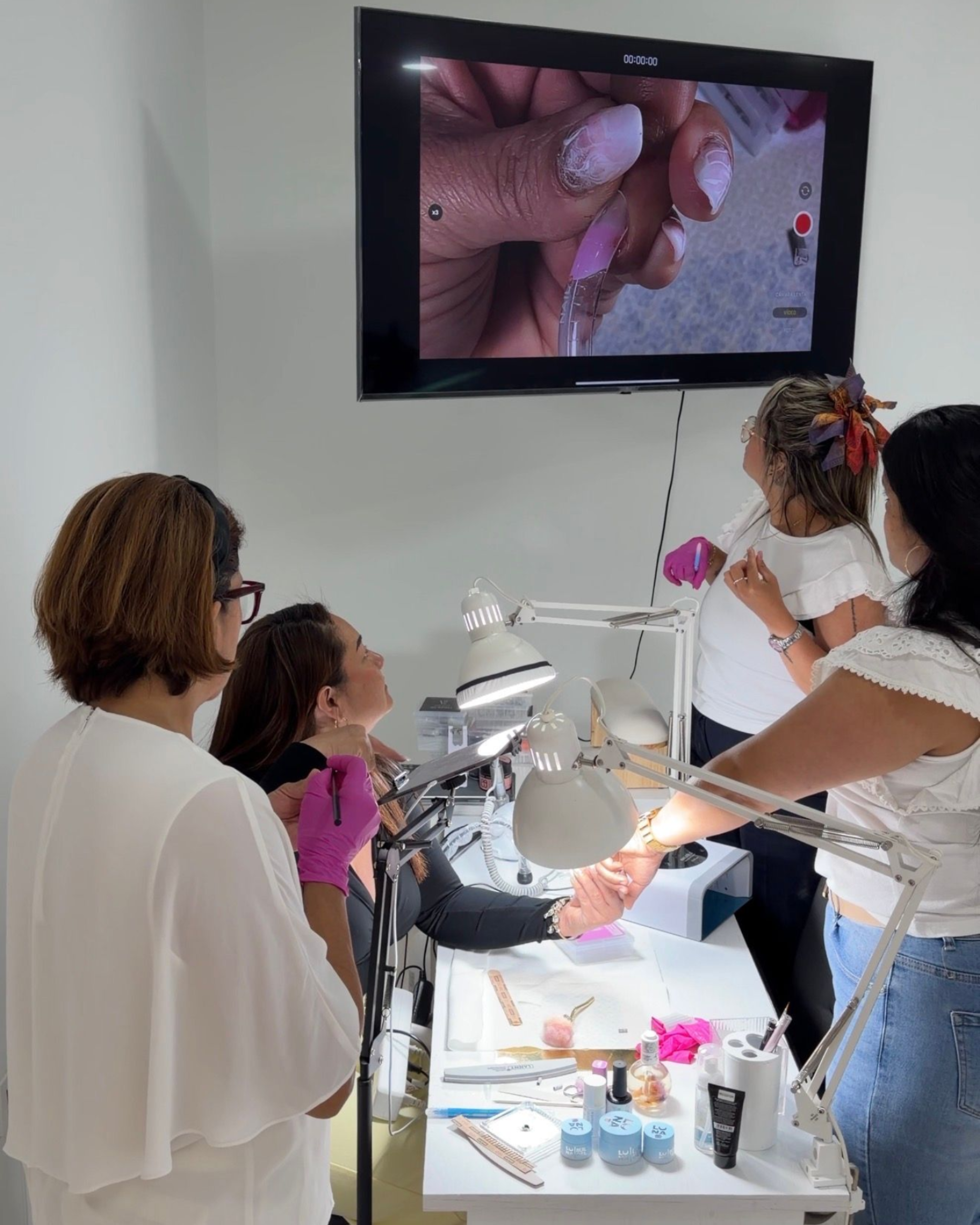Una mujer está sentada en un puesto de manicura mientras otra mujer le aplica el producto, y un hombre permanece de pie cerca grabando con un teléfono inteligente.