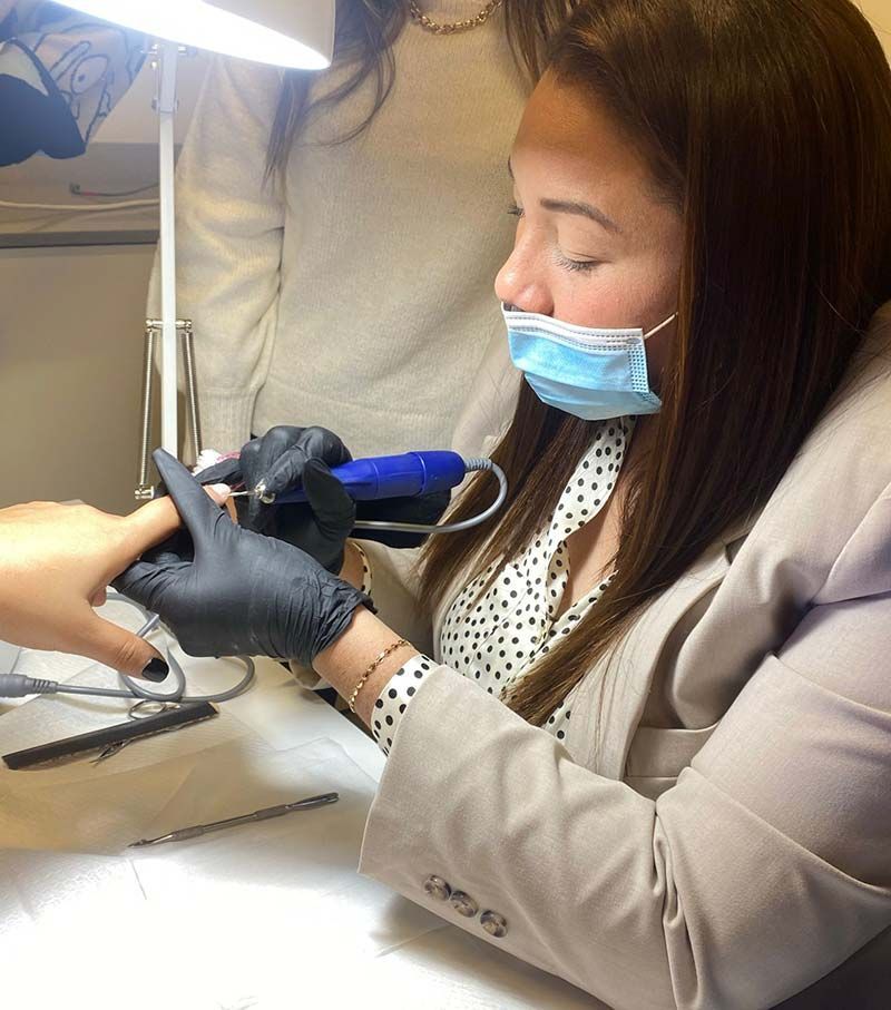 En un salón de manicura, una técnica con mascarilla y guantes negros utiliza una lima de uñas eléctrica en la uña de una clienta.