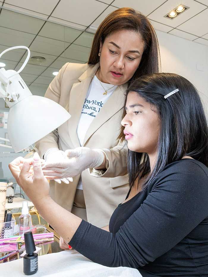 Una profesional de la belleza, vestida con una chaqueta beige y guantes, examina las uñas de una persona con camisa negra en un salón de belleza.