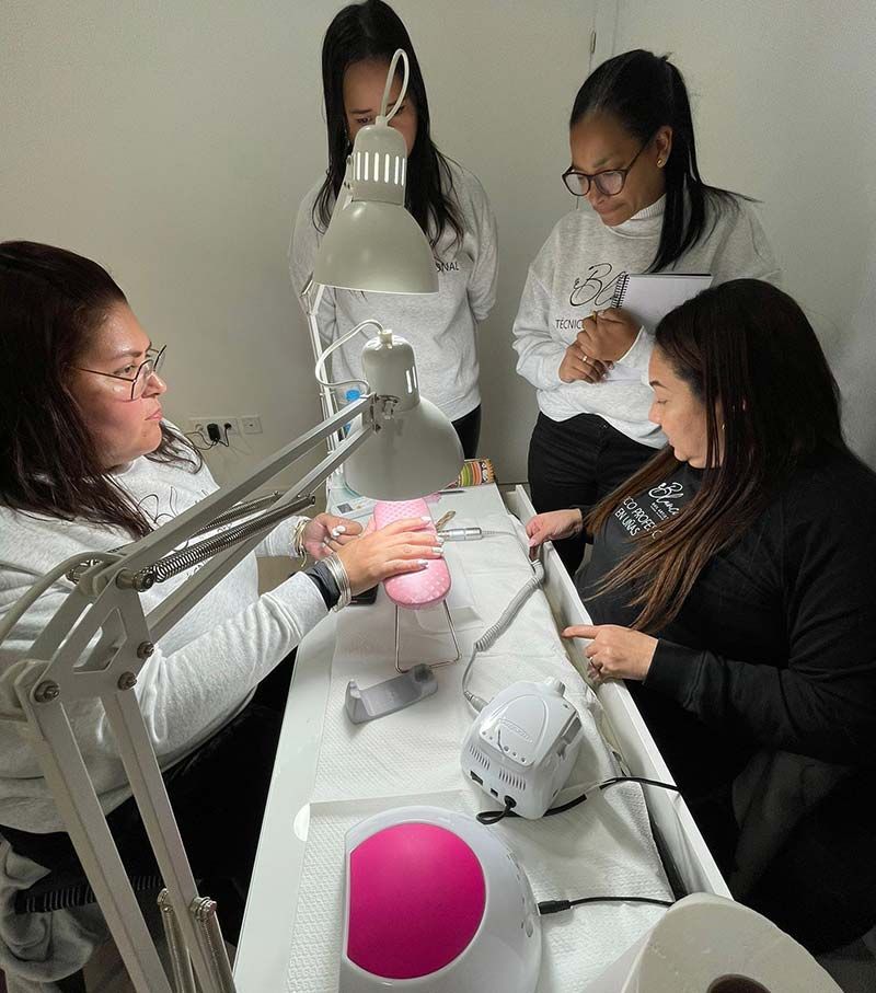 Una técnica de uñas demuestra una técnica de manicura a tres estudiantes en una mesa de entrenamiento equipada con lámparas y otros aparatos.