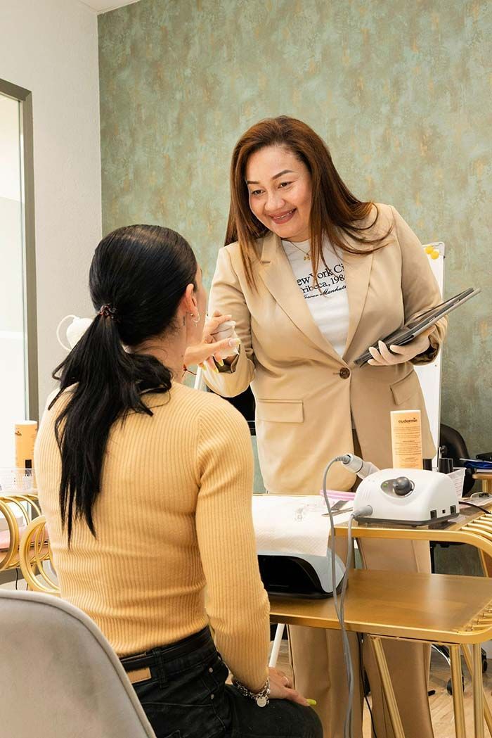 Una persona con un blazer beige está de pie en un puesto de manicura, interactuando con una clienta sentada que lleva una camisa amarilla de manga larga.