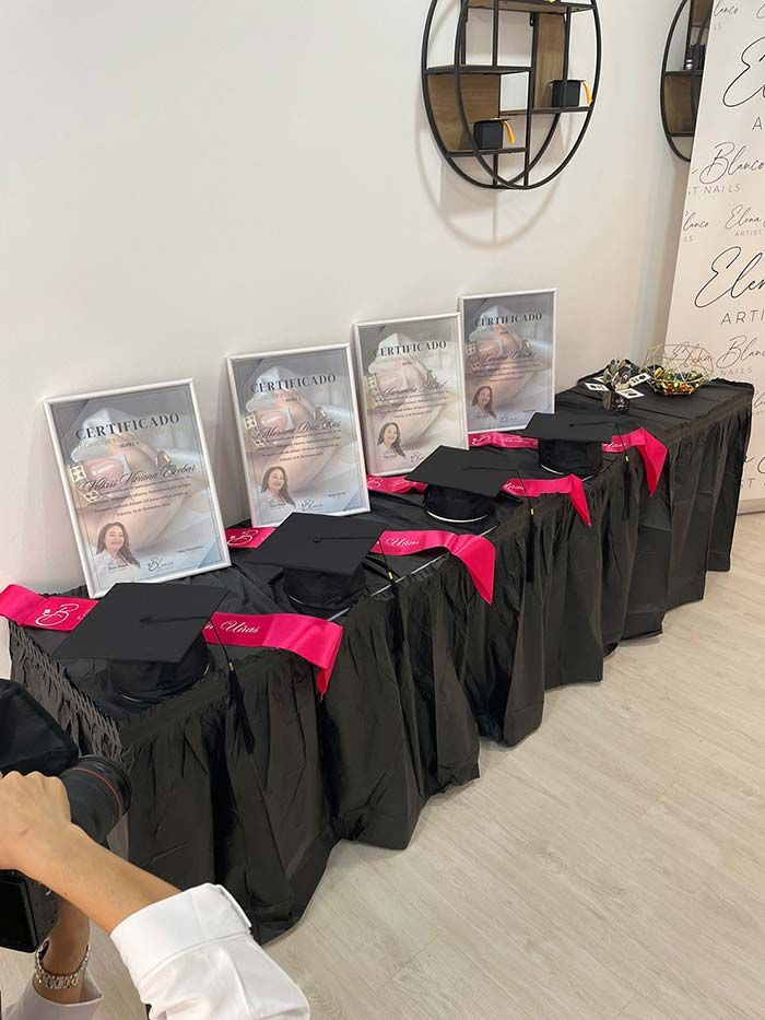 Una hilera de birretes de graduación con bandas rosas reposa sobre una mesa cubierta con un mantel negro, junto a fotografías enmarcadas, en una habitación de tonos claros.