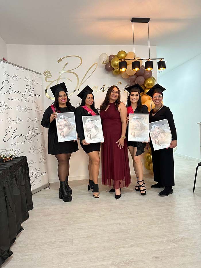 Cuatro graduados con toga y birrete sostienen sus diplomas, de pie junto a una persona vestida con un vestido granate frente a una exhibición de globos dorados.