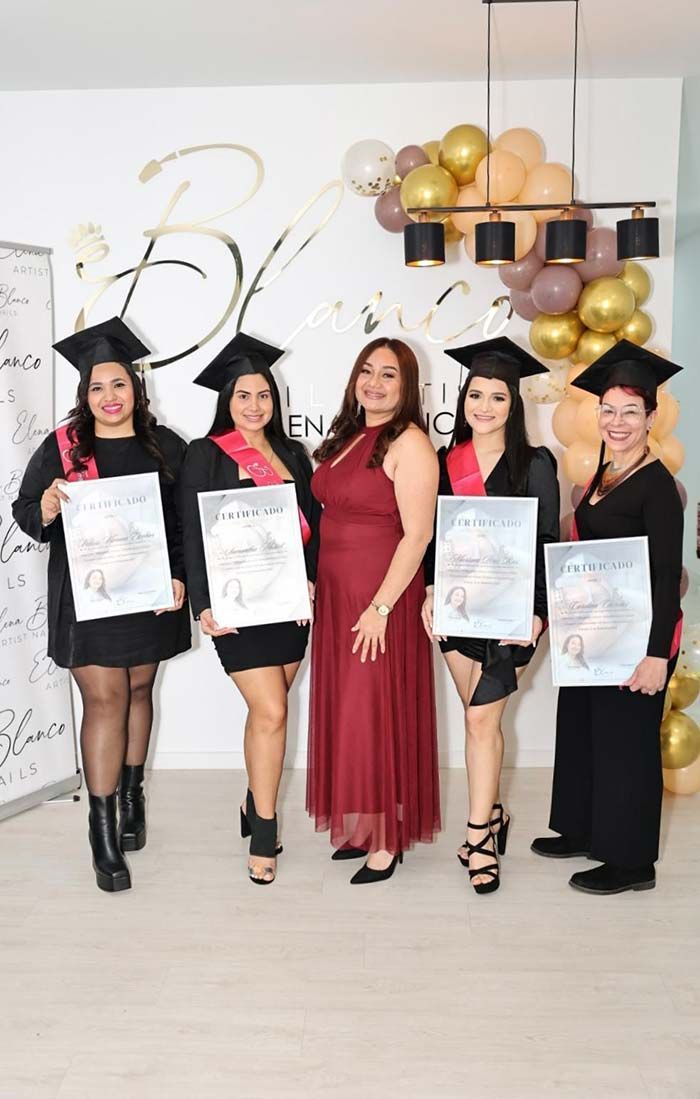 Cinco personas con toga y birrete de graduación están de pie en fila, sonriendo y sosteniendo sus certificados, con globos decorativos detrás.