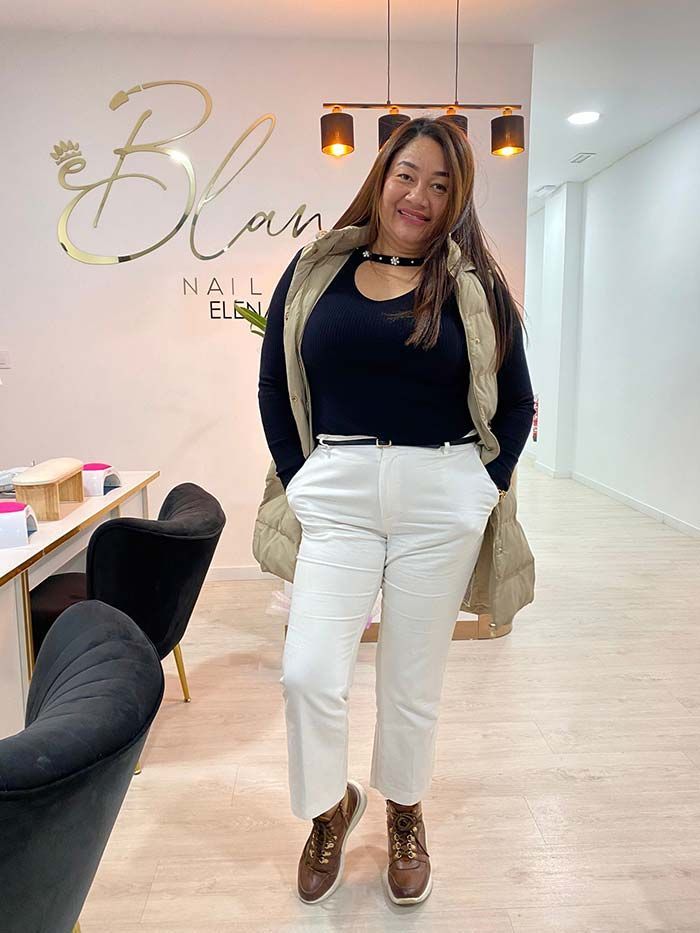 Una persona sonriente, vestida con una blusa negra, pantalones blancos y un chaleco color canela, se encuentra en un salón de manicura muy iluminado.