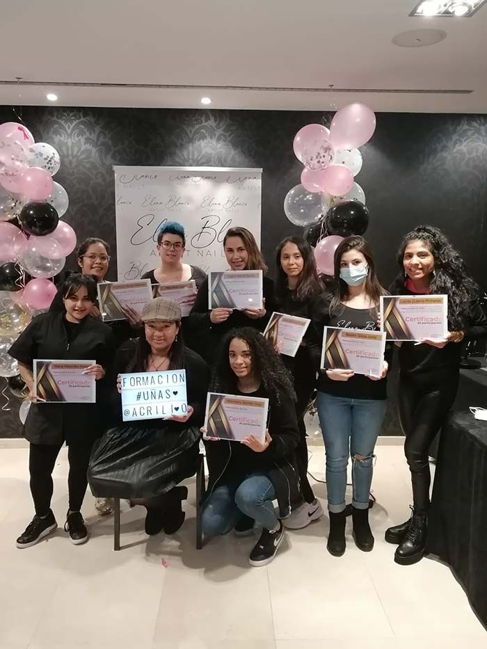 Un grupo de personas sostiene certificados en el interior de un edificio, frente a una pancarta blanca y globos, celebrando un acto de graduación.