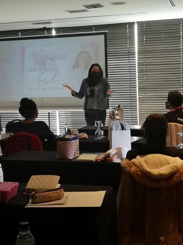 Una persona se encuentra de pie frente a una pantalla de proyección, haciendo gestos hacia el público en una sala de conferencias con grandes persianas.
