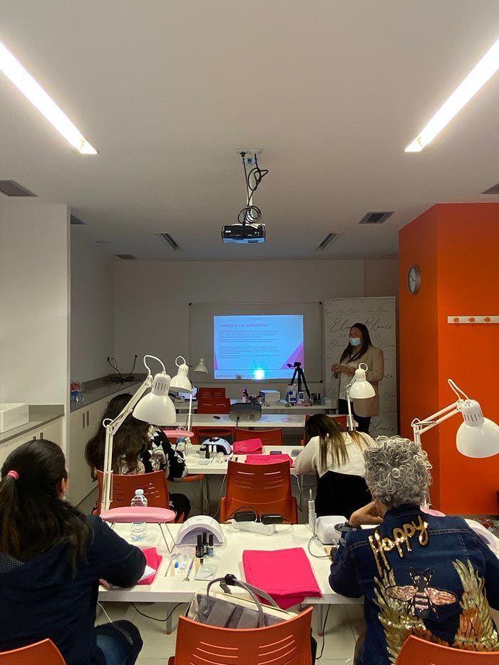 En un aula con pantalla de proyección, se ve a un grupo de estudiantes sentados en pupitres con lámparas durante un taller de belleza.