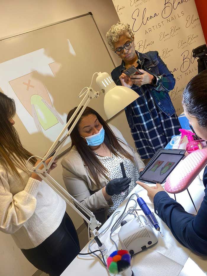 Una técnica de uñas, con mascarilla, trabaja en un escritorio mientras otras dos personas la observan en un aula con una pizarra blanca.