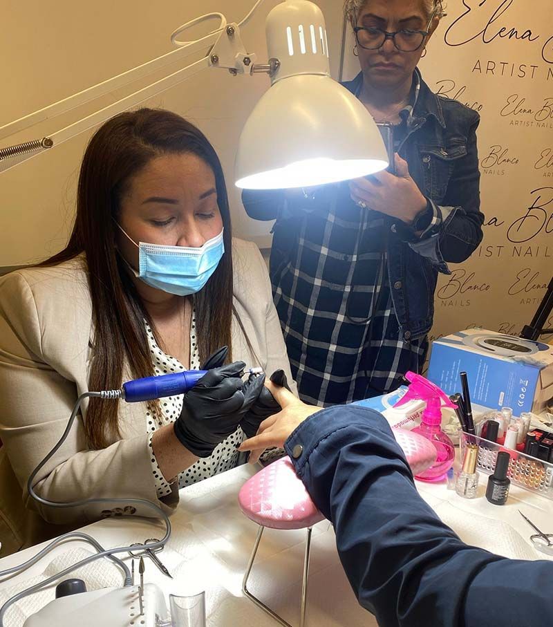 Una profesional trabaja en las uñas de una clienta con una lima eléctrica en un escritorio, mientras una mujer de pie al fondo la observa.