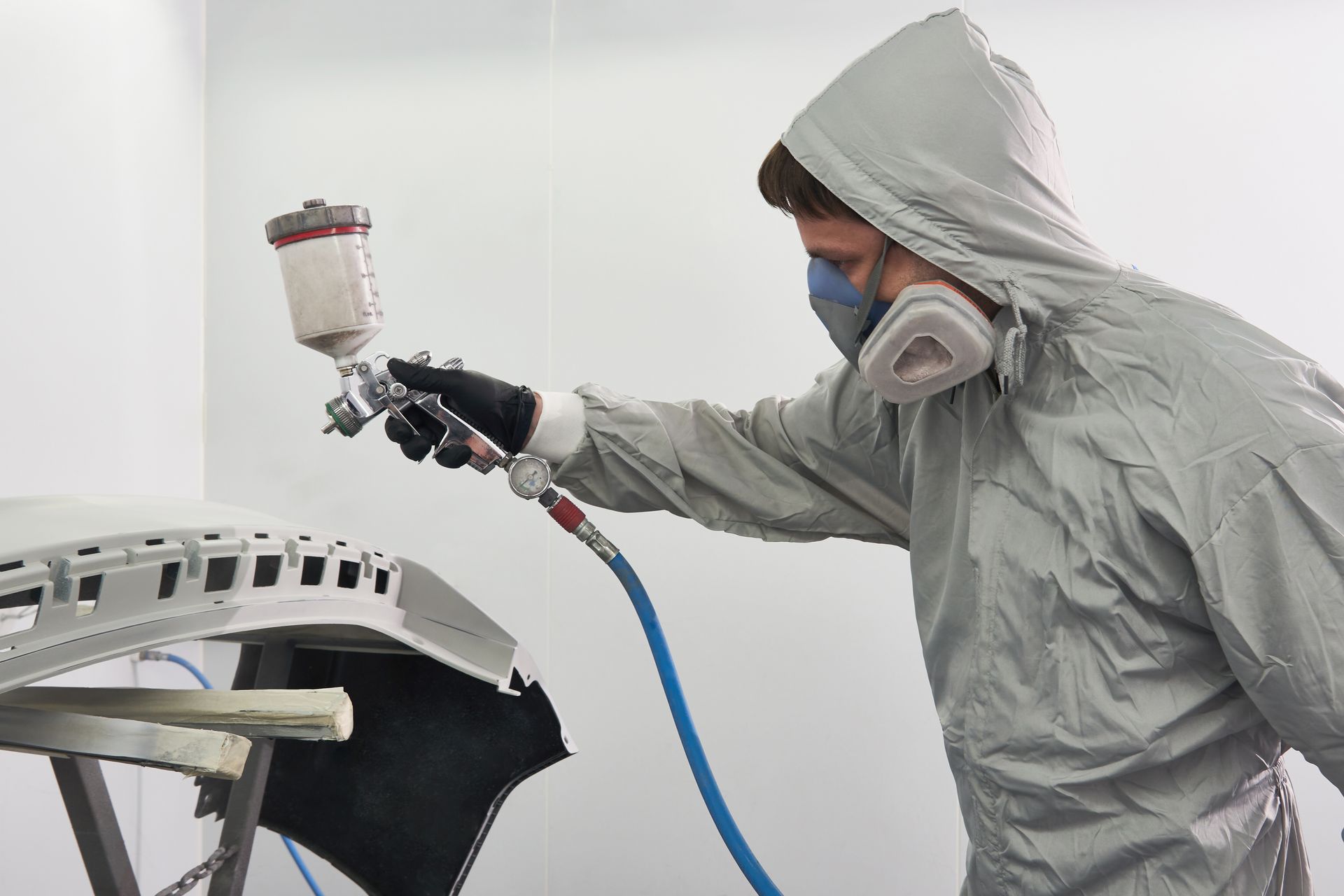 Une personne en combinaison de protection utilise un pulvérisateur de peinture sur un pare-chocs de voiture dans un atelier.