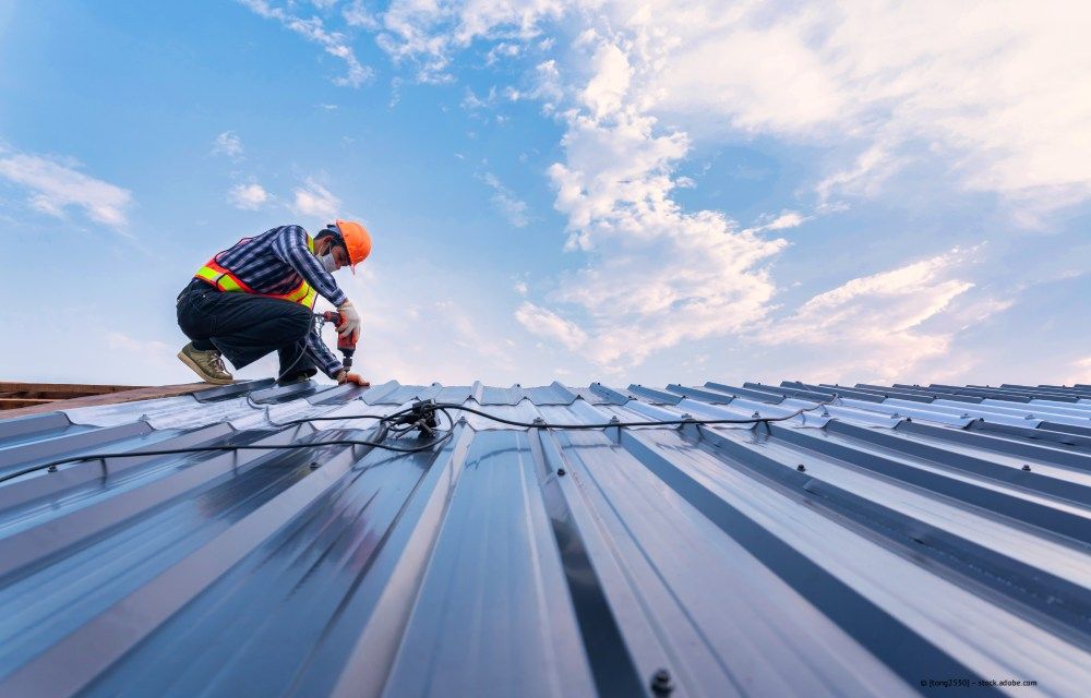 Ein Arbeiter mit orangefarbenem Helm montiert unter blauem Himmel Dachplatten auf einem Metalldach.
