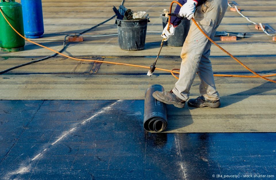 Ein Arbeiter trägt mit einer Rolle eine schwarze, wasserdichte Beschichtung auf ein Flachdach auf.