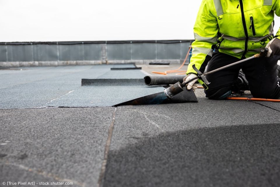 Ein Arbeiter in einer neonfarbenen Jacke brennt Dachmaterial auf einem Flachdach ab.
