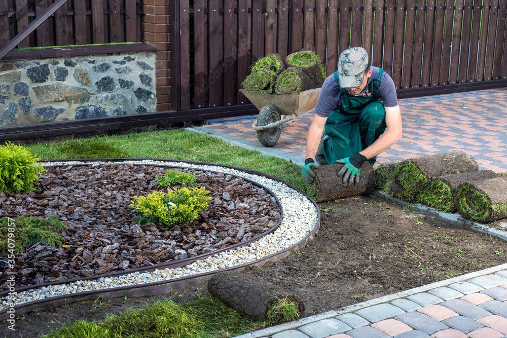 Une personne agenouillée dans un parterre de jardin plante de petits arbustes le long d'une bordure de pierres incurvée.