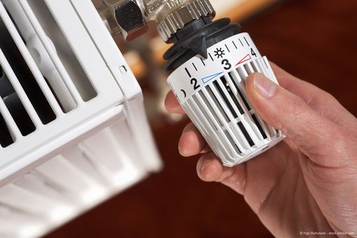 Hand stellt den Thermostatregler eines Heizkörpers an einem weißen Heizkörper in einem Raum von „2“ auf „3“ ein.