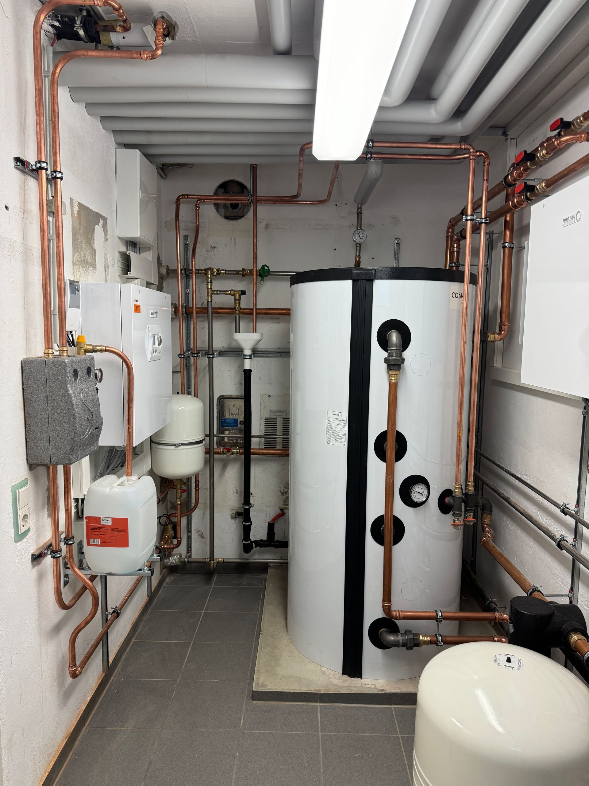 Sanitärsystem in einem Hauswirtschaftsraum: Kupferrohre, weißer Tank, elektrische Komponenten an weißen Wänden, grauer Boden.
