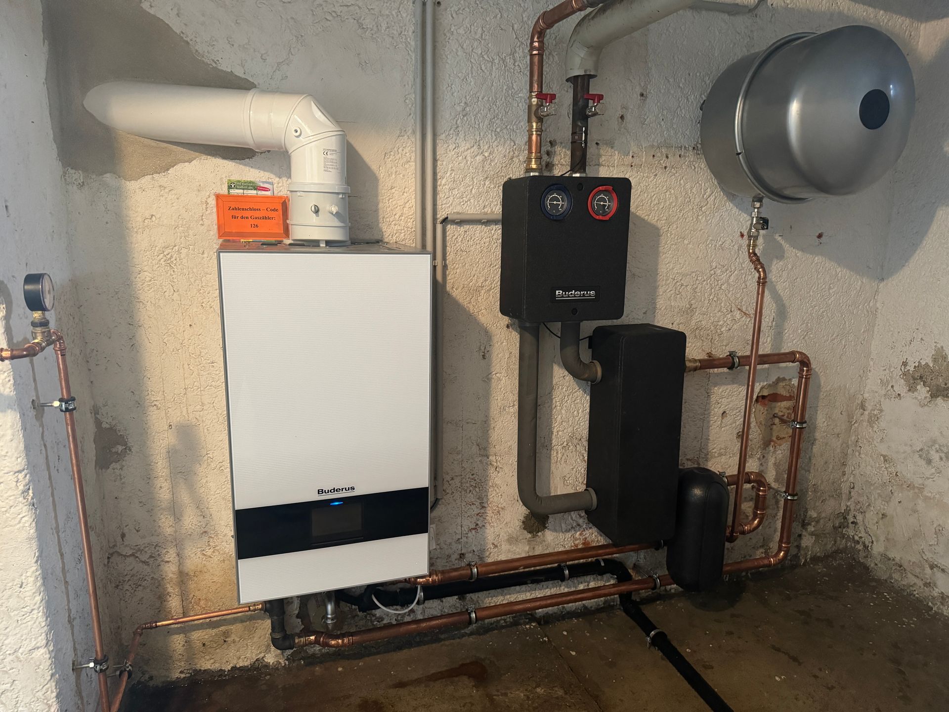 Weißer Ofen mit Kupferrohren, schwarzen Komponenten und einem silbernen Ausdehnungsgefäß in einem Keller.