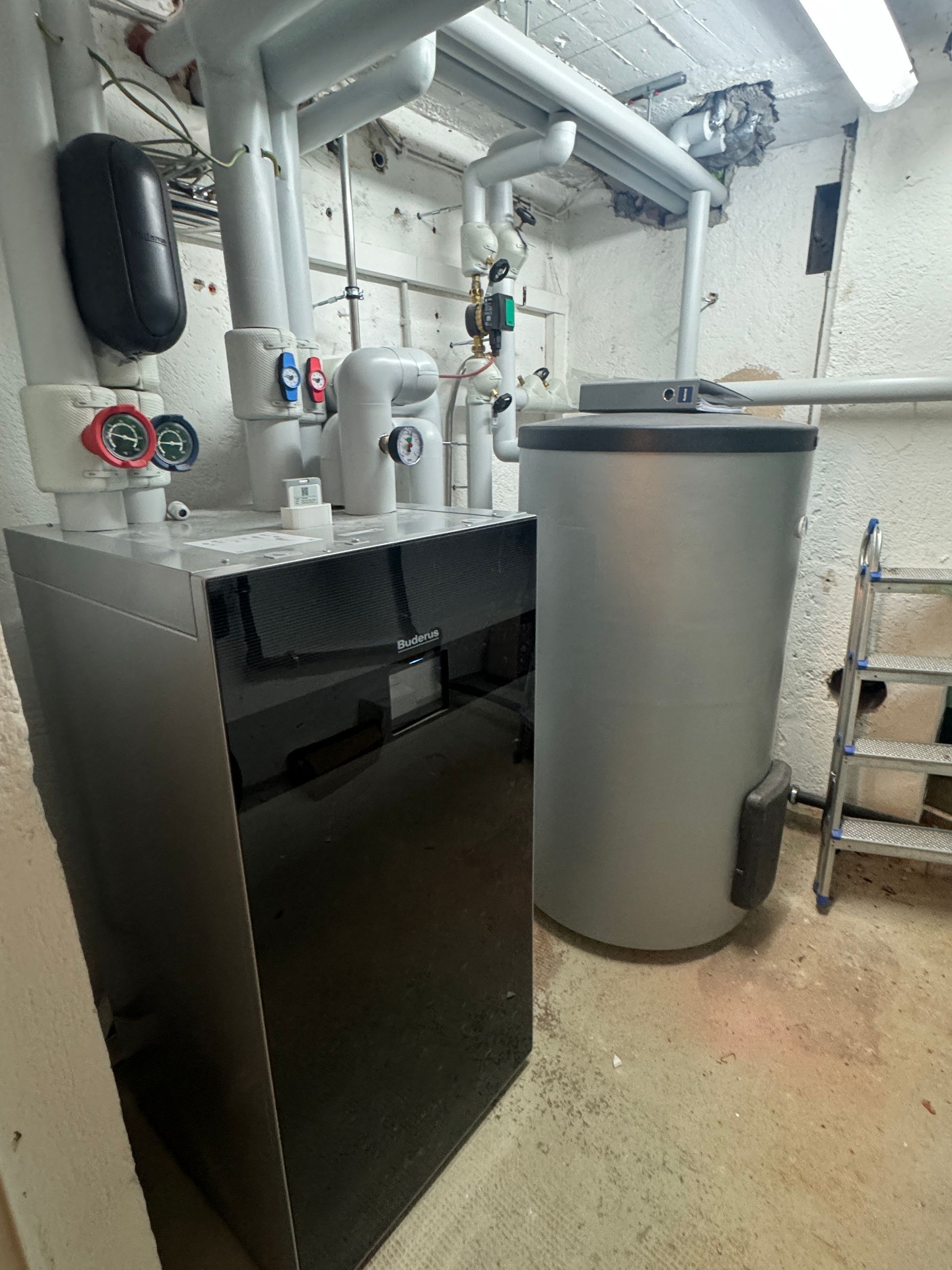 Ein Boiler und ein Warmwasserbereiter in einem Hauswirtschaftsraum mit Rohren und einer kleinen Trittleiter.
