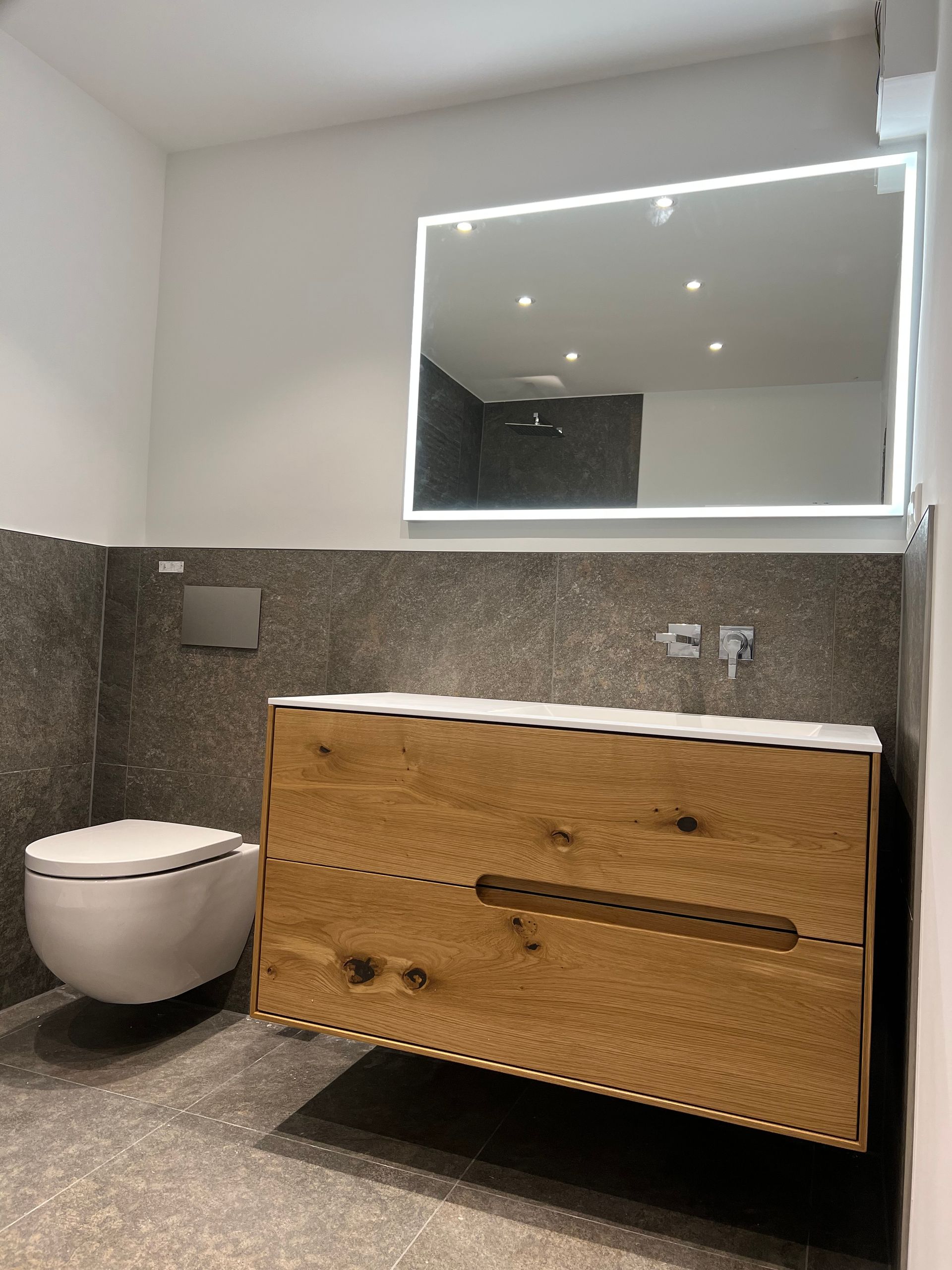 Modernes Badezimmer mit wandmontiertem WC, Holzwaschtisch und beleuchtetem Spiegel. Grau geflieste Wände und Boden.
