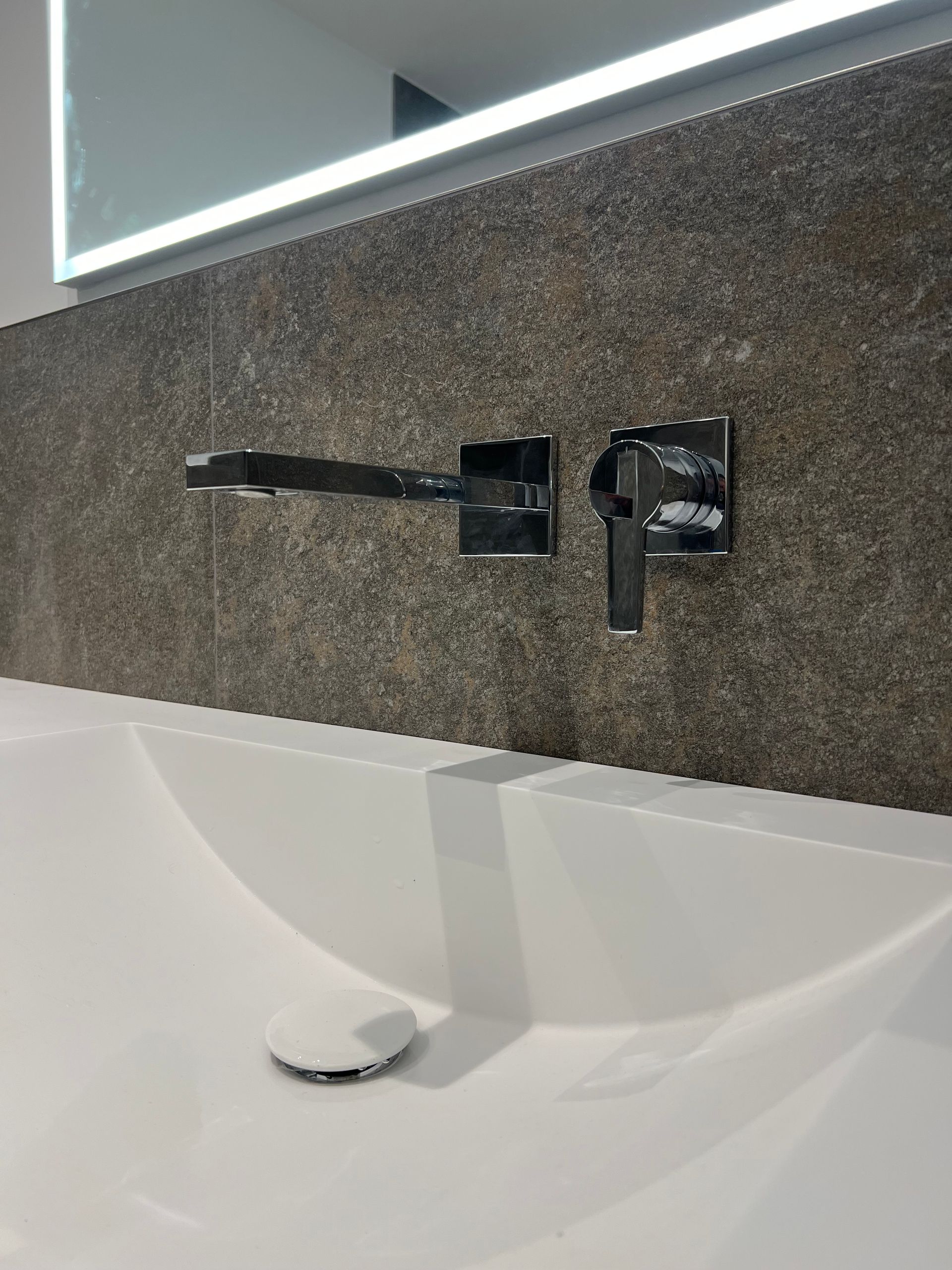 Badezimmerwaschbecken mit an der Wand montiertem Wasserhahn, weißes Waschbecken und Fliesenrückwand mit dunkler Struktur.