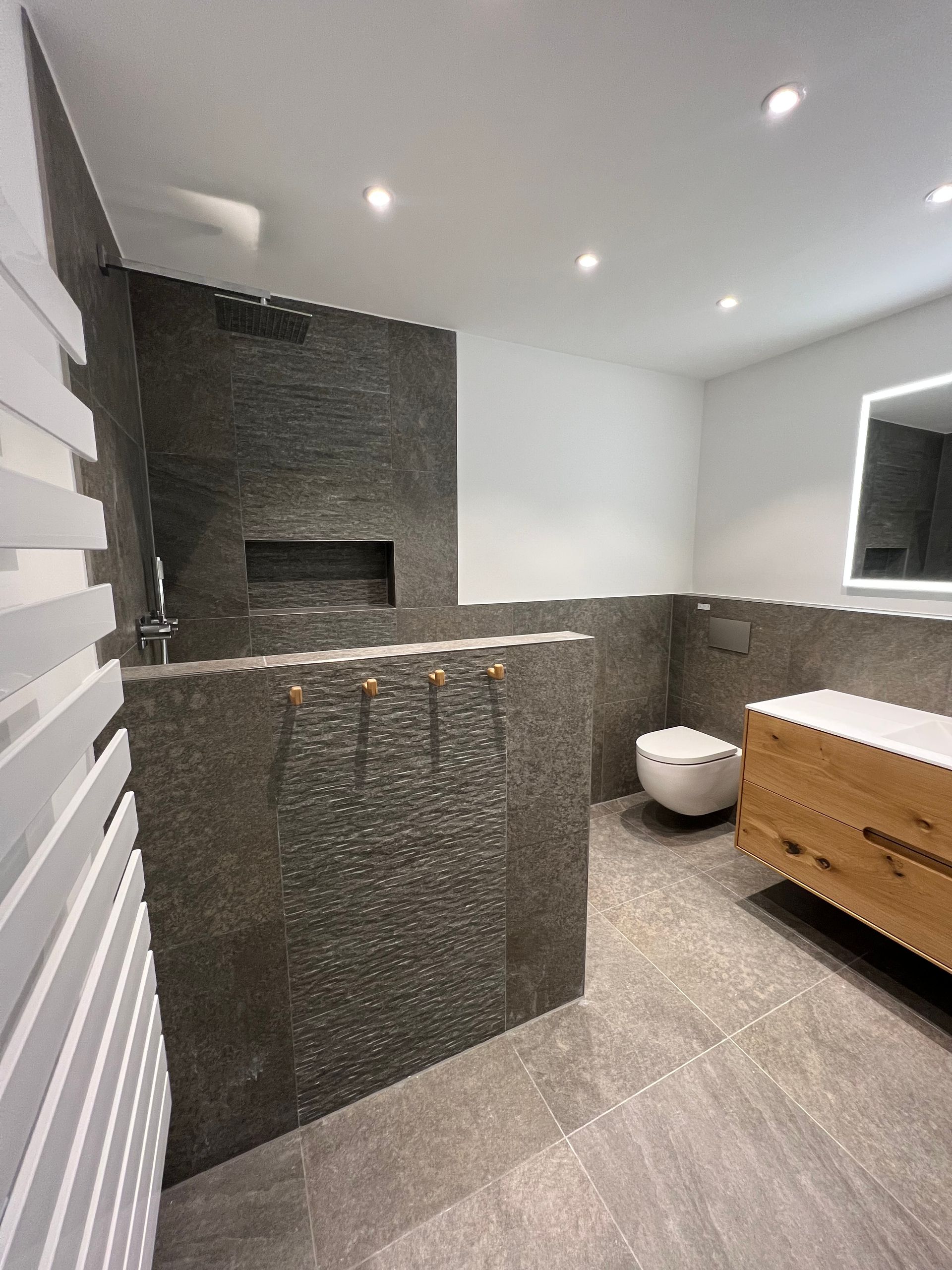 Modernes Badezimmer mit grauen Steinwänden, weißer Toilette, Holzschrank und Einbaudusche.