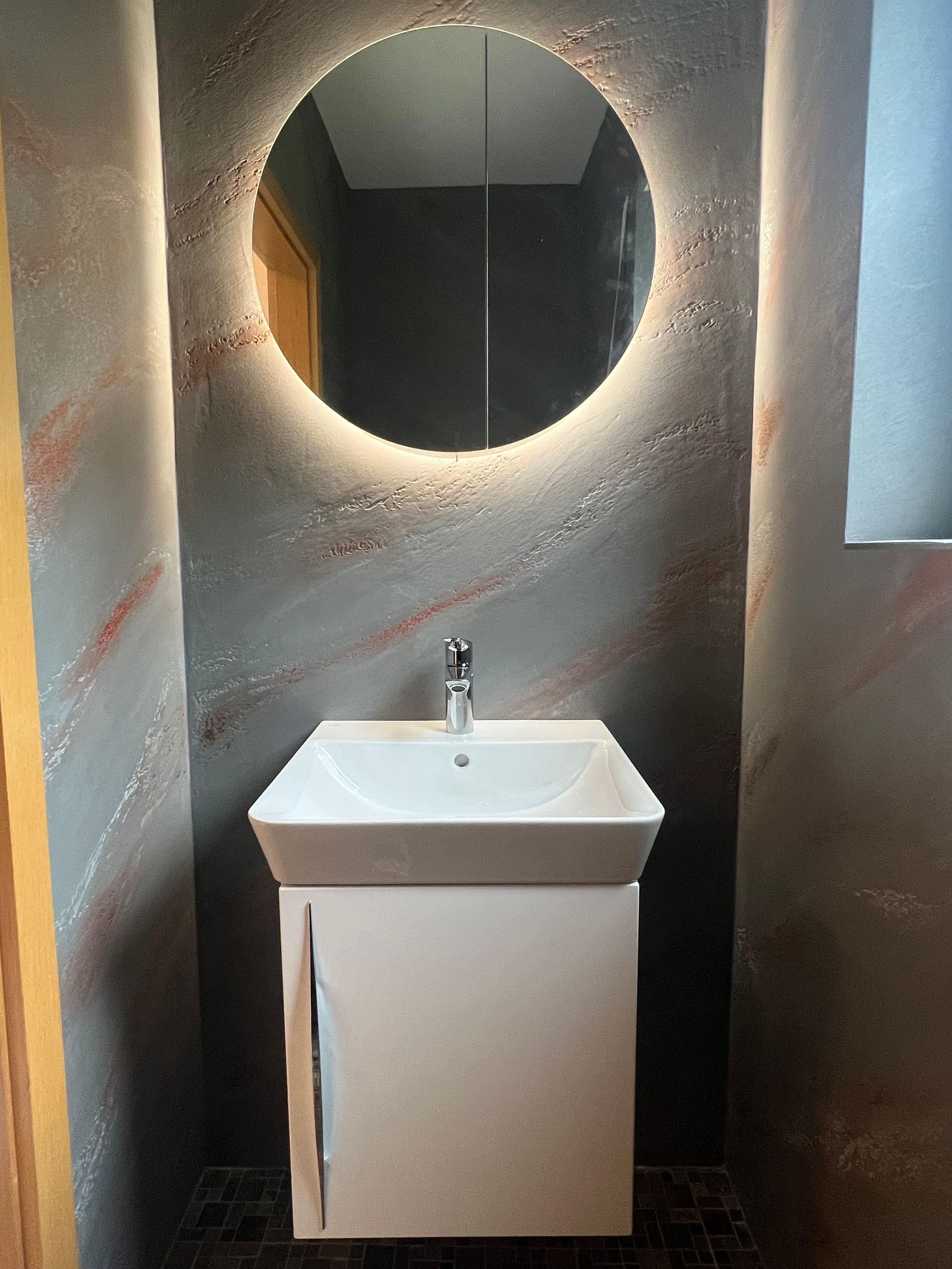 Modernes Badezimmer mit weißem Waschbecken, rundem Spiegel mit LED-Beleuchtung und strukturierten grauen Wänden.