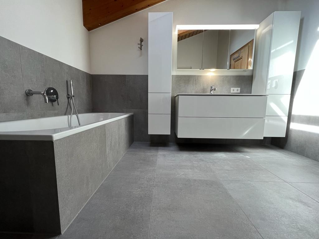 Modernes Badezimmer mit grau gefliesten Wänden und Boden, weißen Schränken und einer Badewanne.