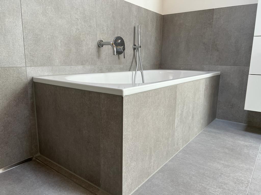 Eingebaute Badewanne mit grau gefliesten Wänden und Boden. Armaturen aus Chrom. Weiße Wanne, neutrale Töne.