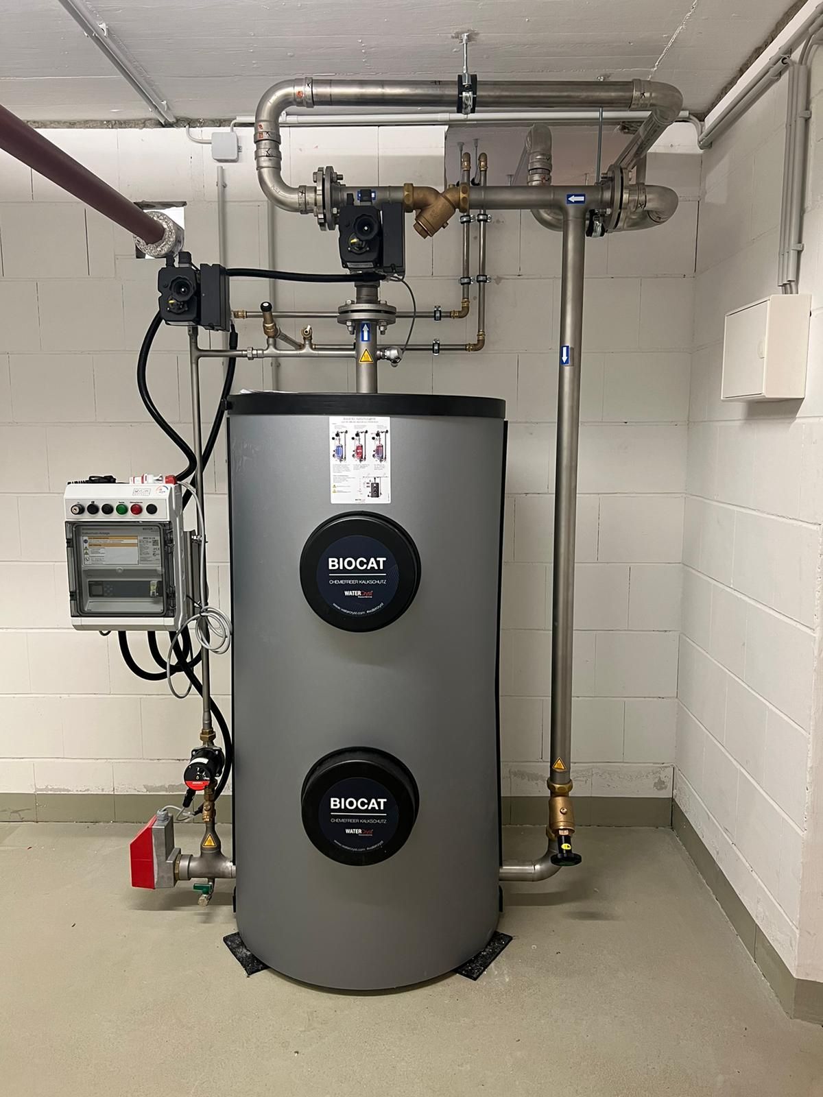 Großer silberner Warmwasserbereiter mit Rohren und Messgeräten in einem Raum mit weißen Wänden.