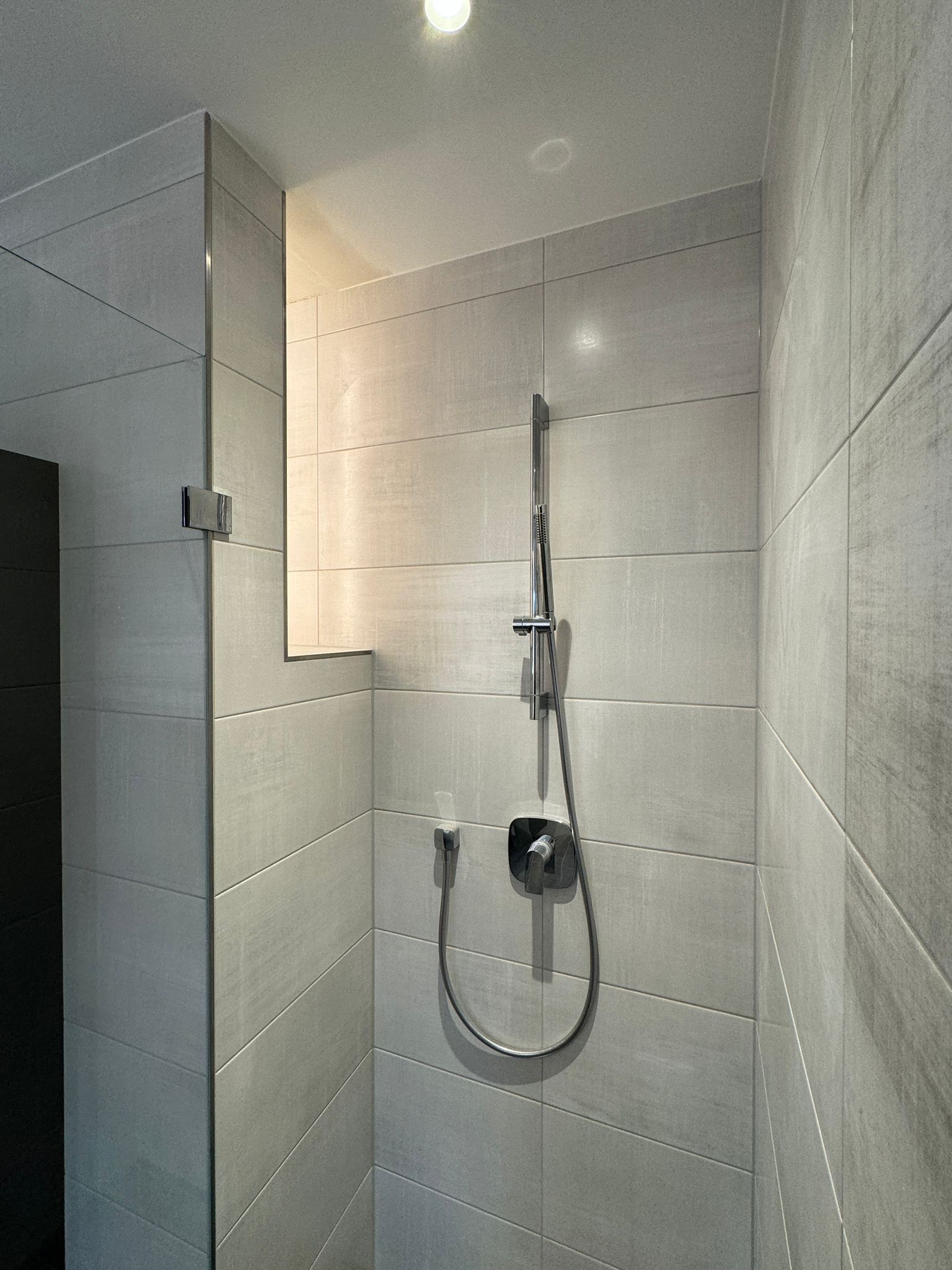 Moderne, grau geflieste Dusche mit Glastür und verchromtem Duschkopf.