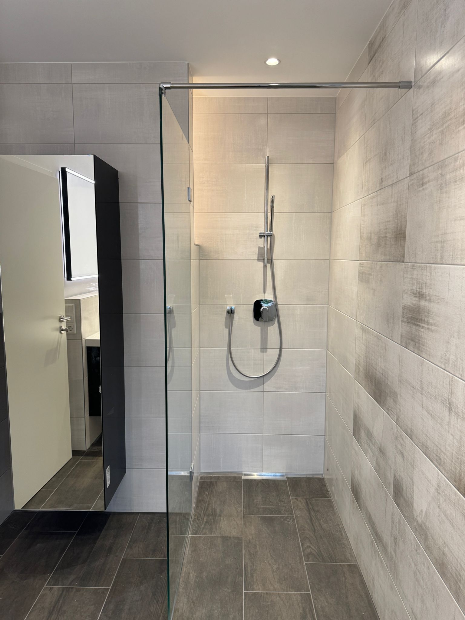 Moderne Dusche mit Glastür, hellgrauen und dunkelgrauen Fliesen. Silberner Duschkopf und Abfluss.