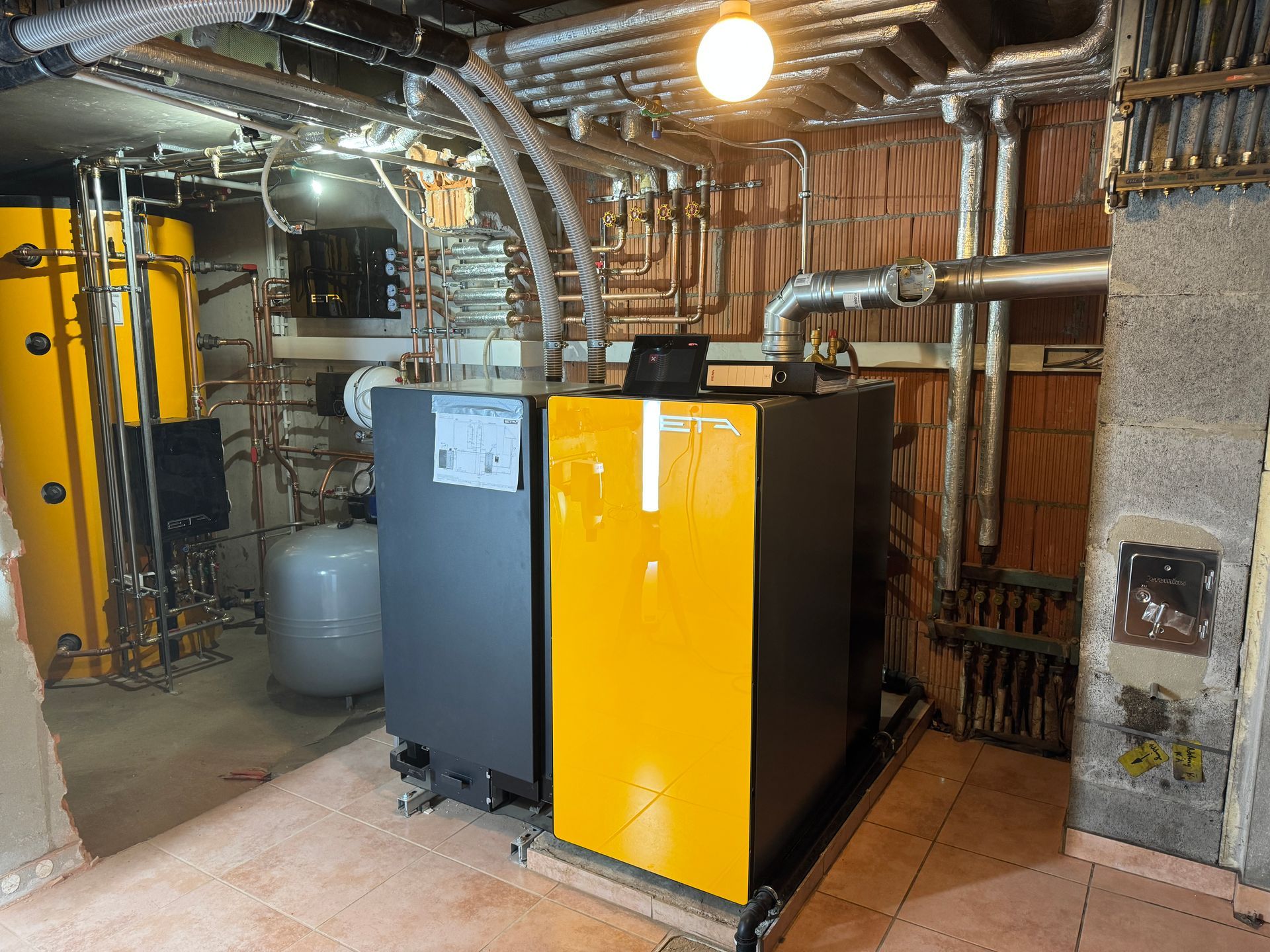 Heizungssystem in einem Keller: gelber und grauer Kessel, Wassertank und Rohre vor Ziegelsteinen und Isolierung.