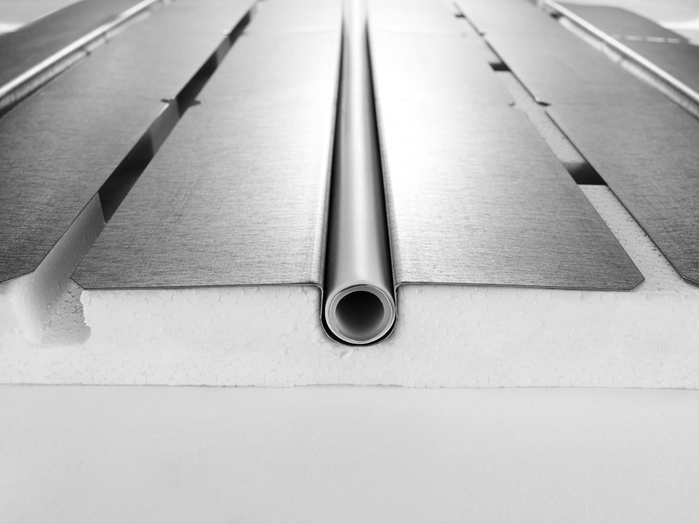 Querschnitt einer Fußbodenheizung: Rohr eingebettet in Isolierung mit metallischen Wärmeverteilungsplatten.