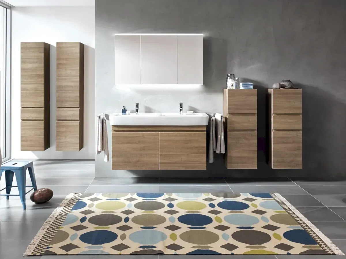 Modernes Badezimmer mit Holzschränken, weißem Waschbecken, grauen Wänden, gemustertem Teppich, blauem Hocker.