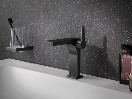 Schwarzer Wasserhahn auf weißem Waschbecken mit Zahnbürstenhalter; glitzernder grauer Wandhintergrund.