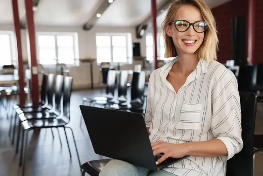 Lächelnde Frau mit Brille, die einen Laptop benutzt, in einem leeren Raum mit Stühlen.