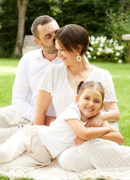 Dreiköpfige Familie, lächelnd auf einer Wiese, der Vater küsst die Mutter auf die Wange.