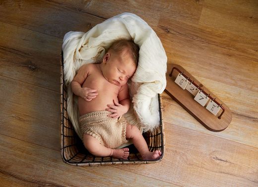 Neugeborenes Baby schläft in einem geflochtenen Korb, der mit einer weißen Decke ausgelegt ist. Blondes Haar, beige Windel, Holzboden.