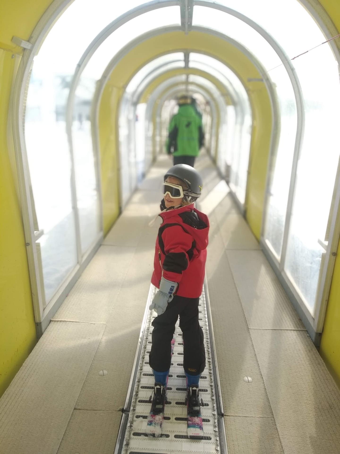 Un niño con chaqueta de esquí roja y gafas está de pie en una cinta transportadora dentro de un túnel, con otra persona delante.
