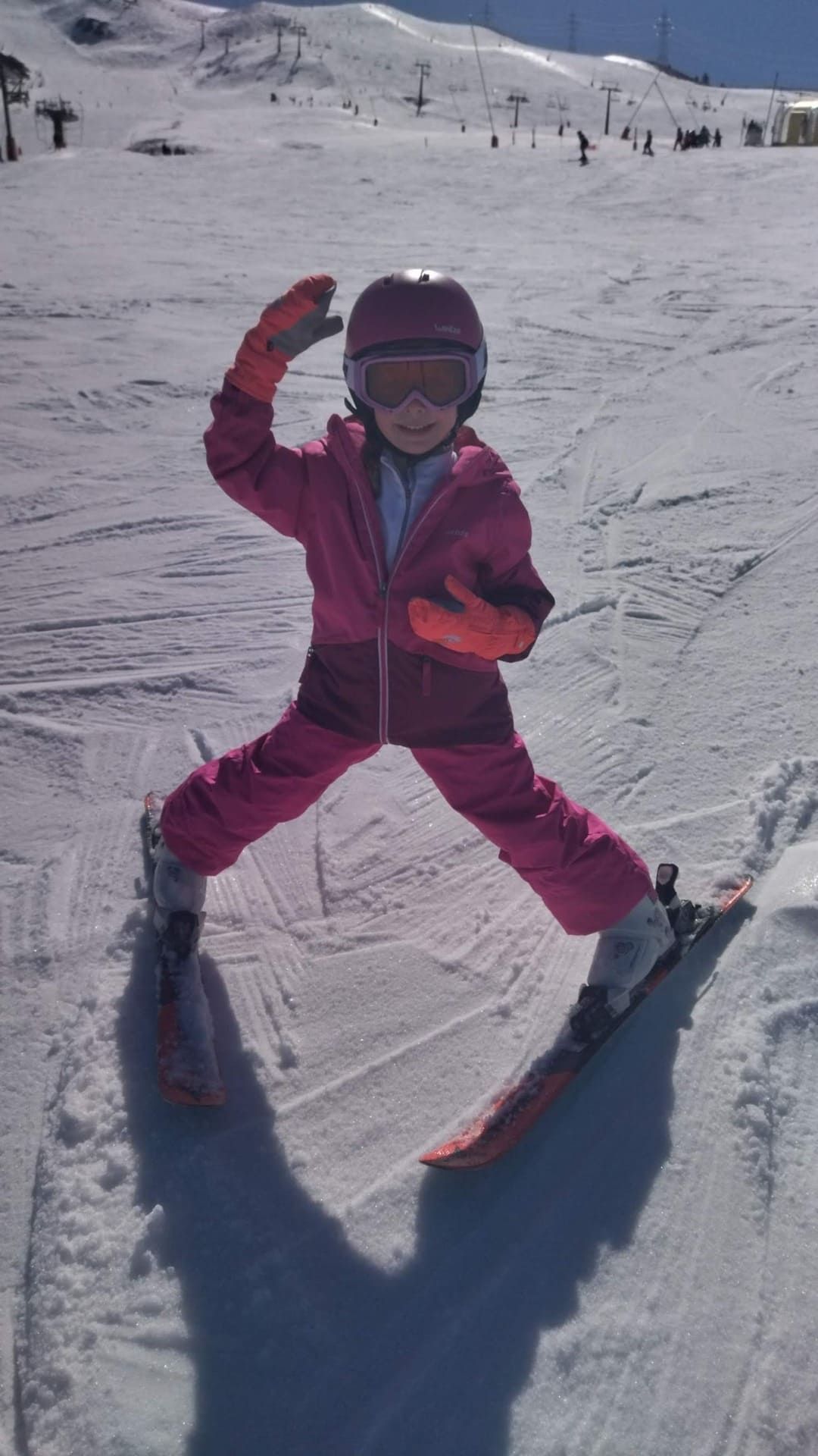 Un niño con ropa de esquí rosa posando sobre unos esquís en una ladera nevada, con un telesilla al fondo.