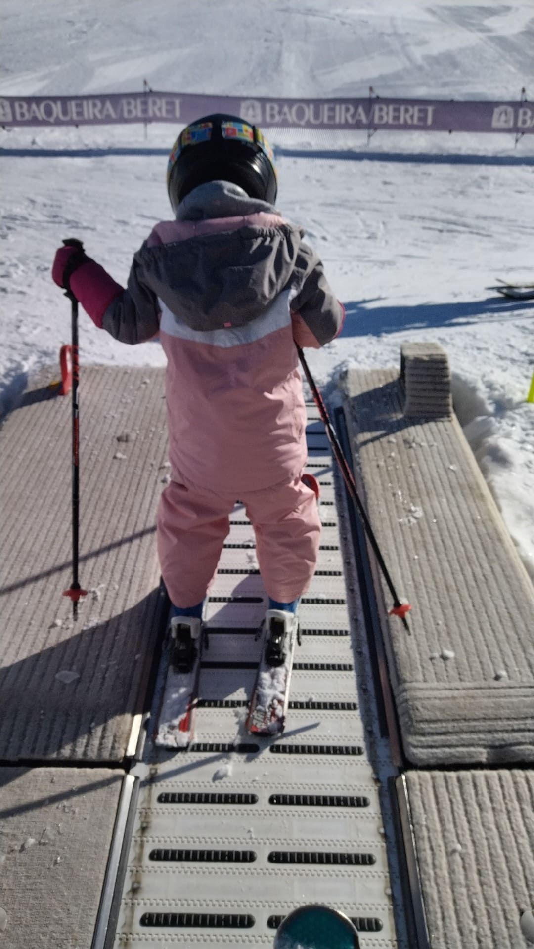 Un niño con ropa de esquí rosa está de pie en la cinta transportadora de un telesquí, sujetando los bastones en una pendiente nevada.