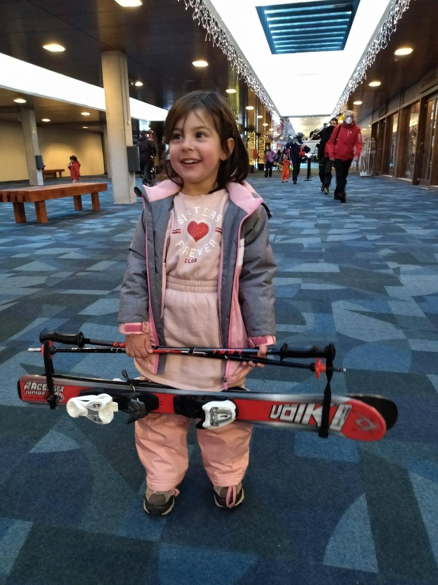 Niño sosteniendo una tabla de snowboard roja en un pasillo interior alfombrado, sonriendo y vestido con ropa de invierno rosa