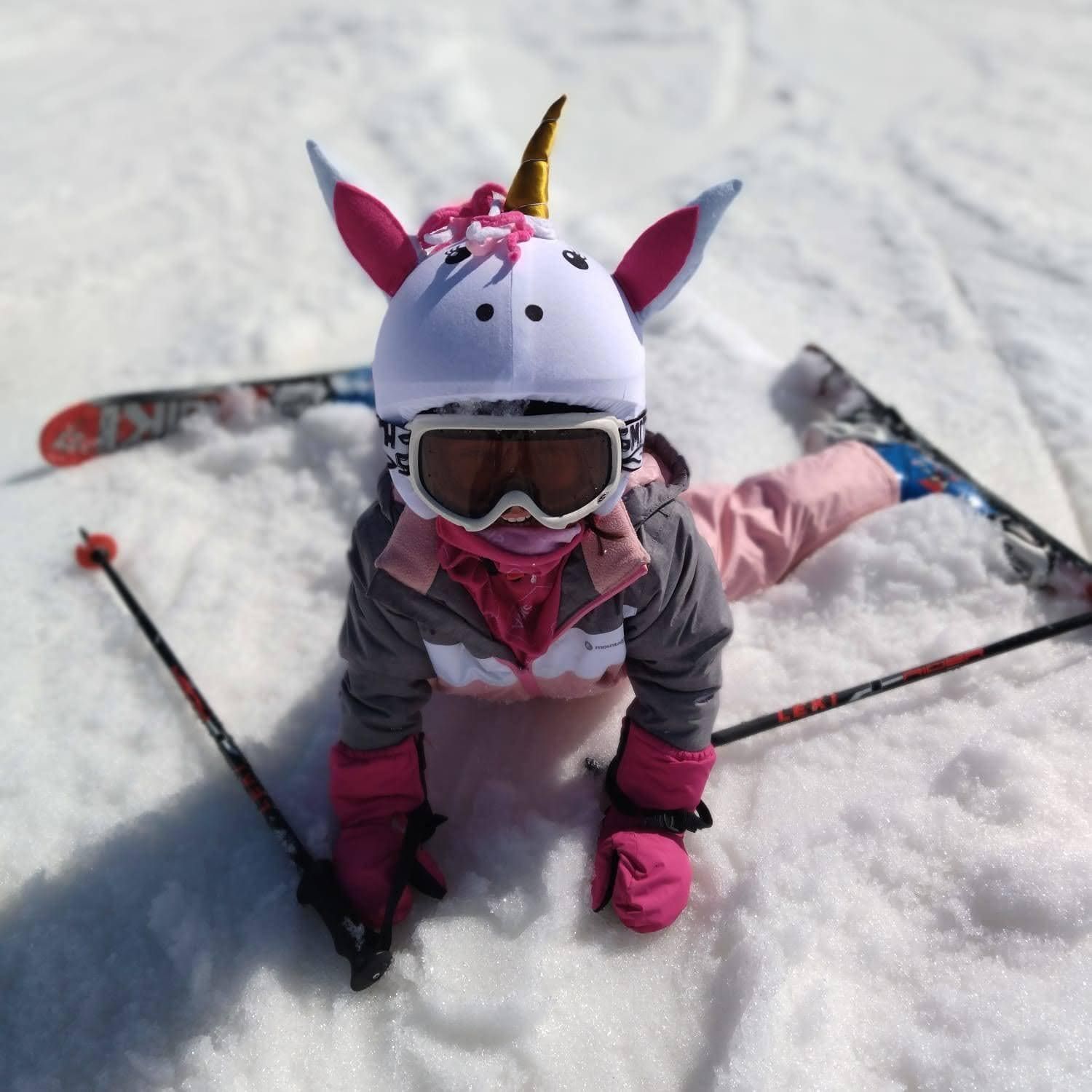 Un niño con casco de esquí de unicornio y gafas se sienta en la nieve con los esquís detrás.