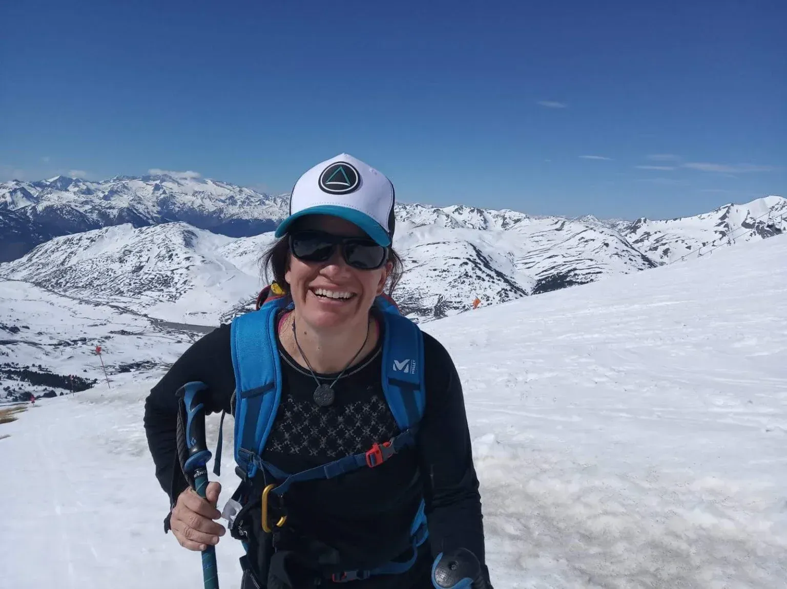 Excursionista sonriente con mochila azul y bastones de trekking en una montaña nevada bajo un cielo azul despejado