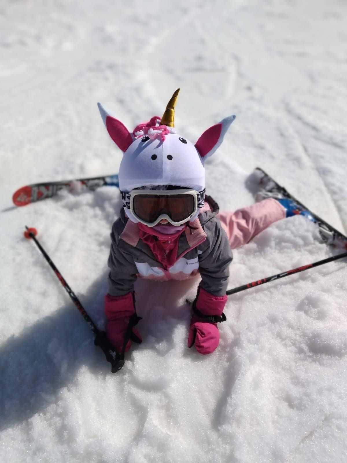 Niño con ropa de esquí rosa sentado en la nieve con esquís y bastones, con casco y gafas con orejas de unicornio