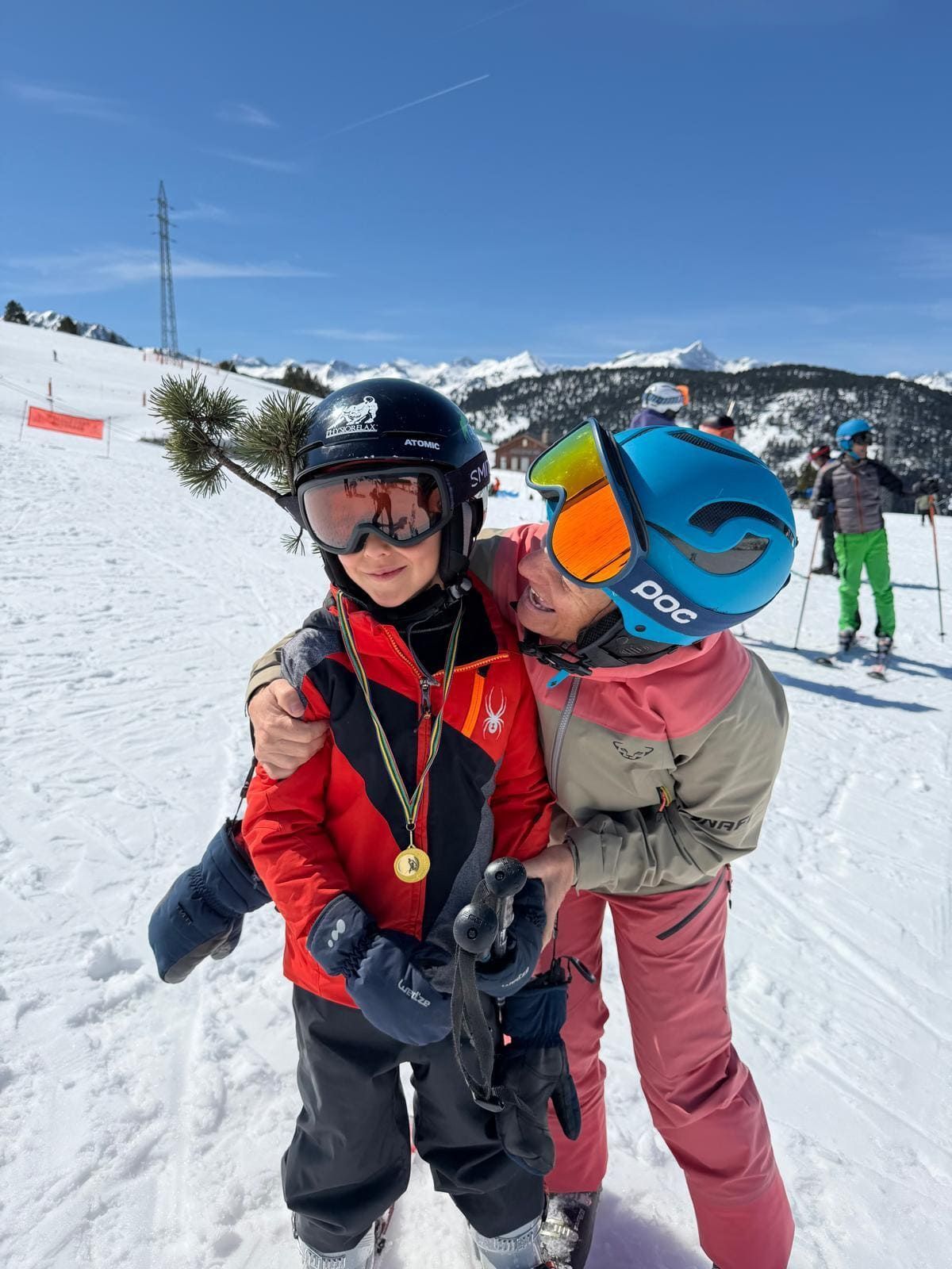 Dos esquiadores abrazándose en una soleada pista nevada; uno lleva una medalla y gafas de esquí